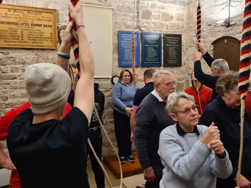 About a dozen people of all ages and genders are crowded round bell ropes in Burghill ringing chamber