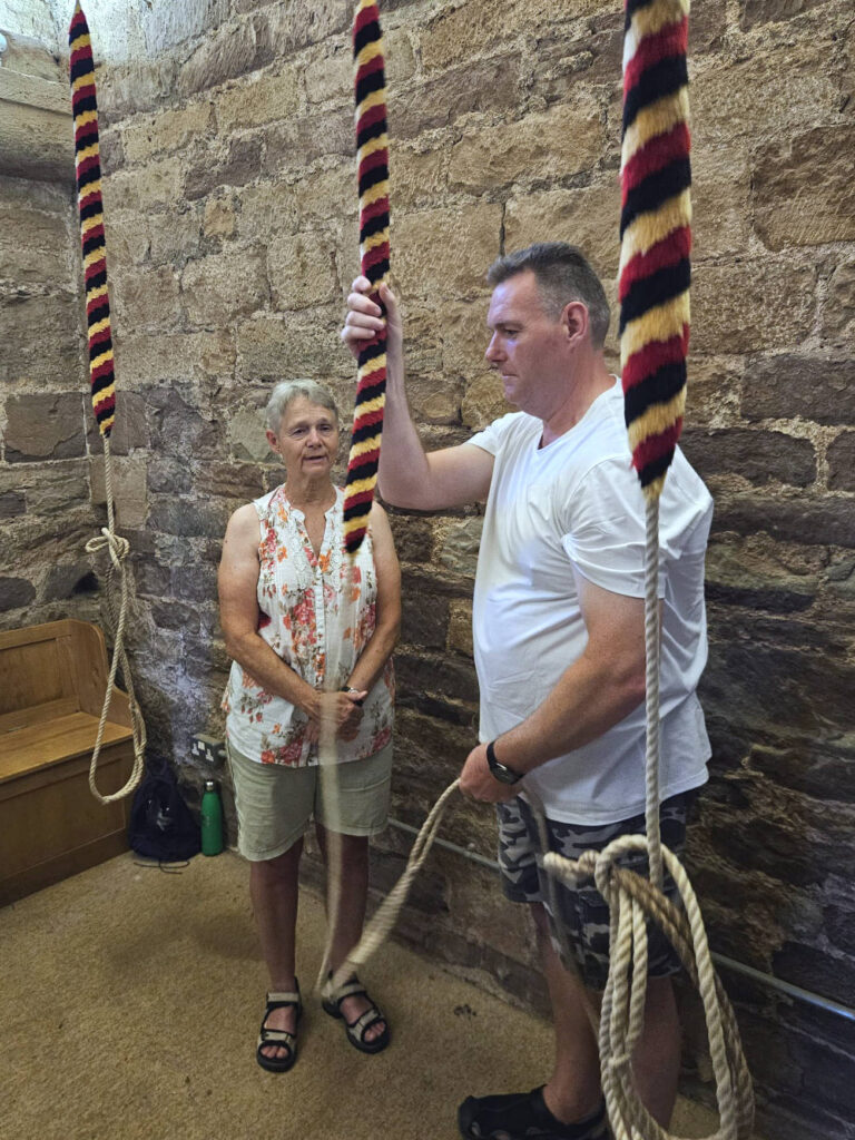 A man in a white t-shirt holds the rope of a bell in his hands, while a female teacher guides him
