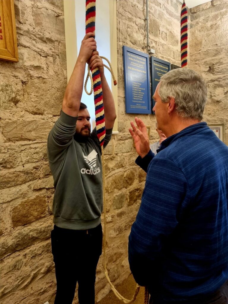 A young man in a green jumper holds the sally of a bell with both hands high above his head, while an older teacher talks to him